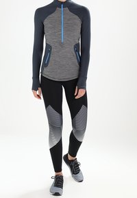 Długi rękaw, top z zamkiem w kolorze szarym z niebieskimi akcentami i kieszeniami; zestawiony z czarnymi legginsami z białymi paskami.