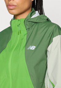 Rompevientos verde y gris claro con cierre frontal, capucha ajustable y logotipo en el pecho. Tejido suave y liviano con un acabado mate.