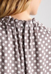 Ryggen af en person iført en taupe bluse med hvide prikker og en lille knaplukning ved halsen.