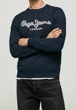 Mężczyzna w granatowej bluzie z kapturem z logo "Pepe Jeans London" na klatce piersiowej, z rękami skrzyżowanymi, ubrany w czarne jeansy.