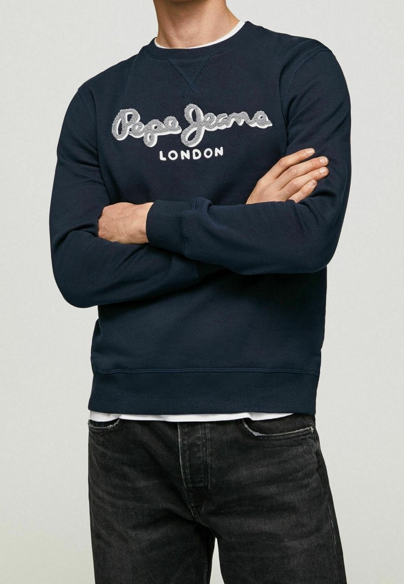 Homme portant un sweat-shirt bleu marine avec le logo « Pepe Jeans London » sur la poitrine, bras croisés, associé à un jean noir.