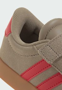 Béžové semišové topánky s červenými pruhmi, s Velcro pásikom. Interiér má vypolstrovaný golier a textúrovanú podšívku. Viditeľná gumová podrážka.