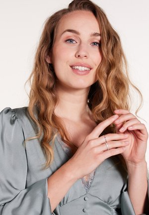 Blouse en soie de couleur gris clair, avec des détails en dentelle et des manches bouffantes ; un modèle aux cheveux ondulés tient délicatement les mains près d'une bague argentée à un doigt.