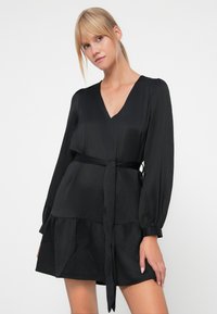 Vestido negro con escote en V, mangas largas y cintura ceñida con cinturón. Presenta una tela texturizada y un dobladillo acampanado con un diseño en capas.
