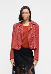 Κόκκινο μπουφάν με σχέδιο και κοντή κοπή, με φαρδύ γιακά, που διαθέτει γεωμετρικά σχέδια σε αντιθετικά χρώματα. Φοράται πάνω από ένα μονόχρωμο κόκκινο τοπ.