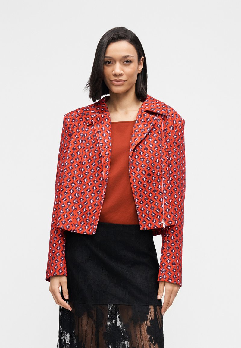 Κόκκινο μπουφάν με σχέδιο και κοντή κοπή, με φαρδύ γιακά, που διαθέτει γεωμετρικά σχέδια σε αντιθετικά χρώματα. Φοράται πάνω από ένα μονόχρωμο κόκκινο τοπ.