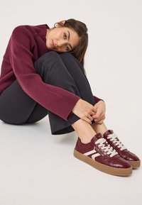 Bordo kožne tenisice s teksturom krokodila, kremastim vezicama i smešim gumenim đonom; uparene s crnim hlačama i rebrastim maroon puloverom.
