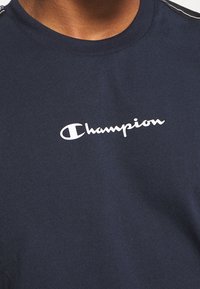 Detailní záběr osoby v námořnickém modrém tričku Champion s bílým logem napsaným na hrudi.