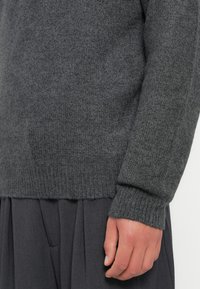 Grijze gebreide trui met een gestructureerd oppervlak, geribde manchetten en een ronde zoom. Draagt over een geplooide donkergrijze kleding voor een gelaagd uiterlijk.