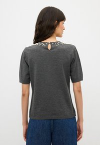 Šedý pletený top s krátkým rukávem, který má klíčovou dírku na zádech a ozdobný límec s perlovými akcenty. Hladká textura, uvolněný střih.