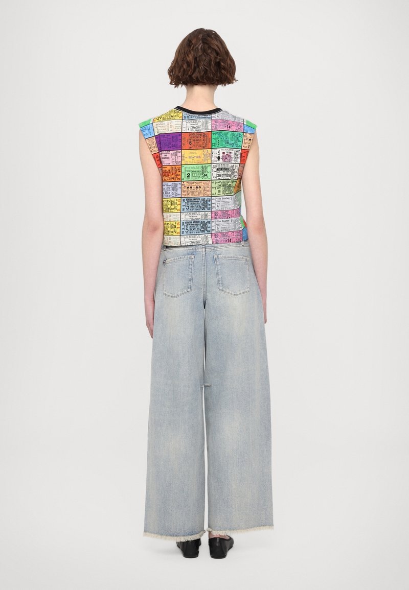 Personne aux cheveux bruns courts portant un haut patchwork multicolore sans manches et un jean bleu clair à jambes larges, debout, de dos.