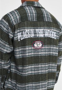 Camicia in flanella a quadri verdi con testo ricamato bianco "PLACING THE WOODS" e una toppa rotonda etichettata "PEGADOR Royalty Club."