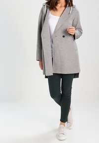 Kvinde iført en lysegrå hættejakke, hvid top, mørkegrønne tætsiddende bukser og hvide sneakers, stående med krydsede ben.