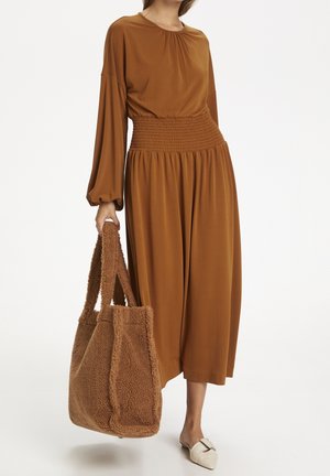 Vestido marrón de manga larga con cintura fruncida y falda fluida. Presenta un escote redondo. La modelo sostiene un bolso de mano peludo de shearling marrón.