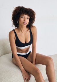 Donna dai capelli ricci che indossa un reggiseno sportivo e culotte neri di Calvin Klein, seduta su una sedia cremosa con texture contro un muro bianco semplice.