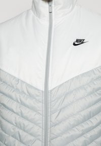 Vit och ljusgrå puffjacka med quiltad textur. Har en hög krage och dragkedjestängning. Svart Nike-logotyp på vänster bröst.