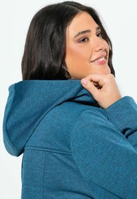 Tummatukka nainen hymyilee pitäen sinisen teksturoidun takin huppua olallaan yksinkertaista taustaa vasten.