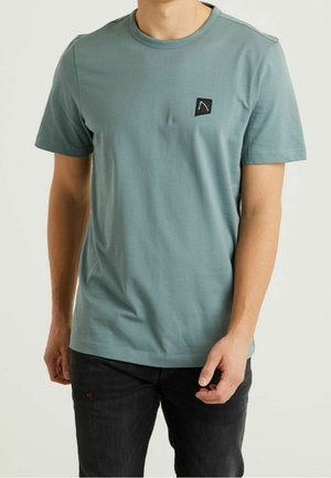 T-shirt en coton bleu sarcelle à manches courtes, doté d'un col rond et d'une petite écusson noir sur le côté gauche de la poitrine. Texture douce et coupe standard.