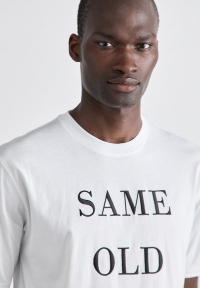 White T-shirt made of cotton with short sleeves, featuring bold black text that reads "SAME OLD" centered on the front. Smooth texture.