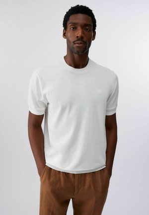 Uomo con capelli corti e ricci, indossa una maglietta bianca a maniche corte semplice e pantaloni marroni, in piedi con le mani nelle tasche su sfondo bianco.