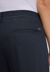 Zbliżenie na kieszeń granatowych, strukturalnych spodni z etykietą w kolorze czarnym i częściowym widokiem osoby noszącej białą koszulę.