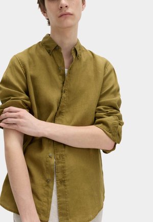 Junge Person in olivgrünem Hemd mit hochgekrempelten Ärmeln, die einen Arm vor einfachem, hellem Hintergrund anpasst.