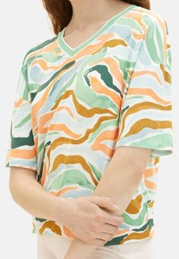 Femme vêtue d'un t-shirt ample à manches courtes et col en V avec des rayures ondulées abstraites en vert, orange, marron et bleu clair.