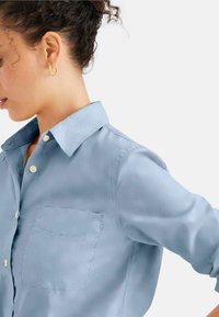 Camicia blu chiaro in tessuto morbido, con colletto, taschino sul petto e bottoni chiari. Capelli dell'utente acconciati a mosso.