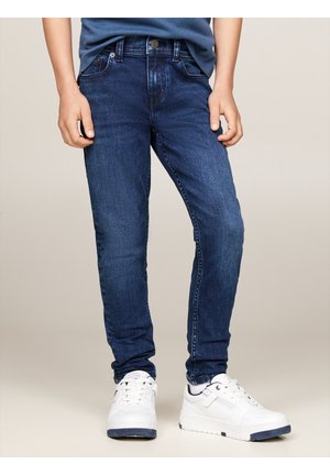 Enfant portant un jean bleu et des baskets blanches, debout, les mains détendues le long du corps, sur un fond uni.