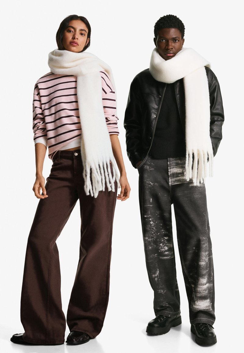 Hvid uld tørklæde hænger om en models hals; stribet lyserød og hvid sweater. Mørkebrune bredde bukser og sorte sko. Metallisk pynt på bukserne.