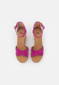 Růžové semišové klínové sandály s překříženými pásky, nastavitelným páskem kolem kotníku a světle hnědou polstrovanou stélkou. Uvnitř je vytištěné logo UGG.