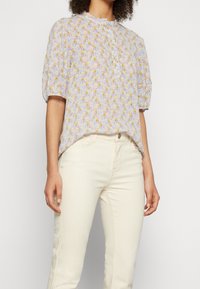 Blusa de manga corta en púrpura claro con un patrón floral en amarillo y verde. Presenta un cuello redondo y un cierre frontal de botones.