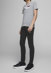 Graues Baumwoll-T-Shirt mit "JACK & JONES" Logo in Rot und Blau, kombiniert mit schwarzen Slim-Fit-Hosen und schwarzen Sneakers mit weißen Sohlen.