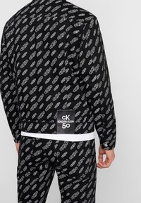 Svart jacka och byxor med ett upprepat vitt "CK50" mönster. Jackan har en etikett på baksidan med "CALVIN KLEIN"-logotyp.