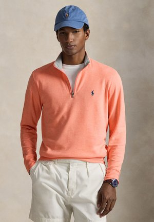 Coral-colored pullover with a quarter-zip design, gray collar lining, and a small blue logo on the chest. Worn with white shorts and a blue cap.