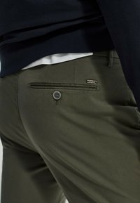 Ingrandimento di una persona che indossa pantaloni verde oliva con tasca posteriore con bottone e parte di una camicia bianca infilata sotto un top scuro.