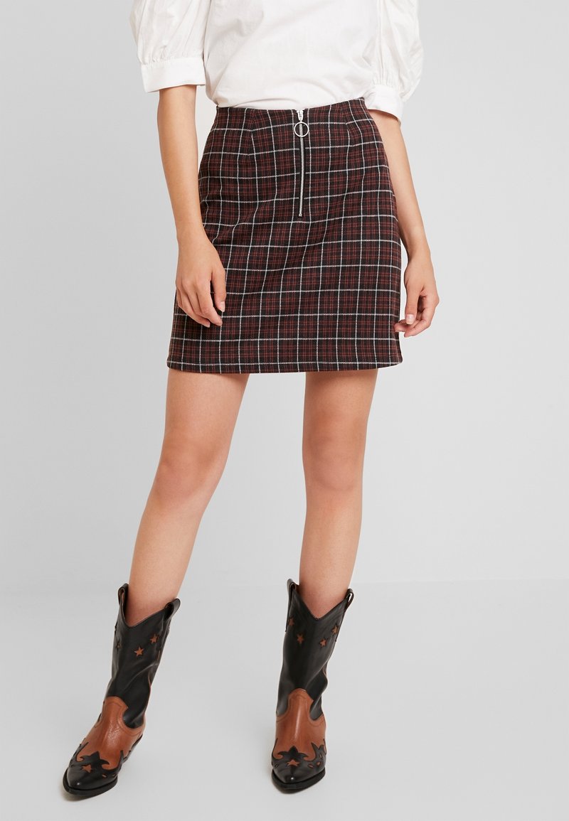 Vrouw in een donkerrode geruite mini-jurk met hoge taille, met een rits aan de voorkant en zwarte cowboylaarzen met bruine accenten en stervormige uitsparingen.