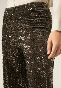 Zwarte broeken versierd met pailletten, met een strakke textuur. Kenmerkt zich door soepel materiaal met een aansluitende snit en een lichte glans. Close-up van de taille en hand.