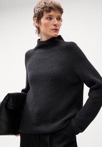 Pletena crna vestica s visokim ovratnikom, rebrastom teksturom i dugim rukavima. Model drži crnu kožnu torbu. Jednostavan, minimalistički dizajn.