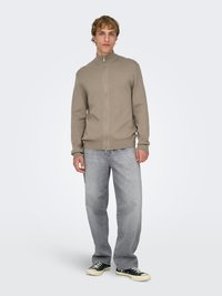 Pull beige à fermeture éclair avec un col montant, associé à un jean droit gris clair et à des baskets basses noires. Texture douce, design décontracté.