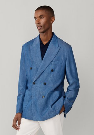 Hombre con blazer cruzado azul claro sobre camisa oscura y pantalones blancos, de pie frente a un fondo gris liso.