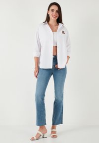 White button-up shirt with short sleeves and a small embroidered bear on the chest, paired with light blue flared jeans and white heeled sandals.