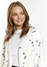 Cardigan bianco texturizzato con accenti di paillettes multicolori, indossato sopra una maglietta bianca semplice. Presenta una vestibilità rilassata e bordi frangiati.
