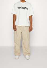 Vit kortärmad t-shirt med svart graffitistil-logotyp, kombinerad med beiga lösa byxor och vita sneakers.