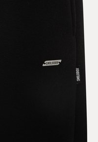 Sorte stretchjoggingbukser med en tekstureret overflade, forsynet med et sølvfarvet "SMILODOX" logo-patch og en sideetiket.