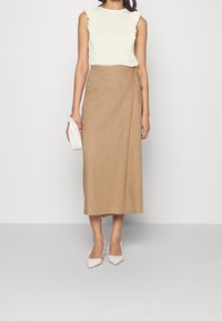 Žena na sobě má bílou bezrukávovou blůzu s volány, béžovou midi sukni s překrytím, bílé lodičky se špičatou špičkou a drží bílou clutch kabelku.
