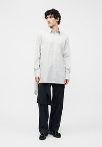 Junge Person mit lockigem Haar, die ein übergroßes, hell gestreiftes Hemd und asymmetrische, dunkel gestreifte Nadelstreifenhosen mit schwarzen Schuhen trägt.