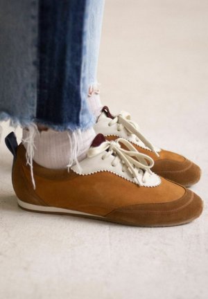 Scarpe in suede color sabbia con dettagli in pelle bianca, di forma arrotondata e con lacci. Jeans blu scoloriti e calzini bianchi con righe leggere visibili.