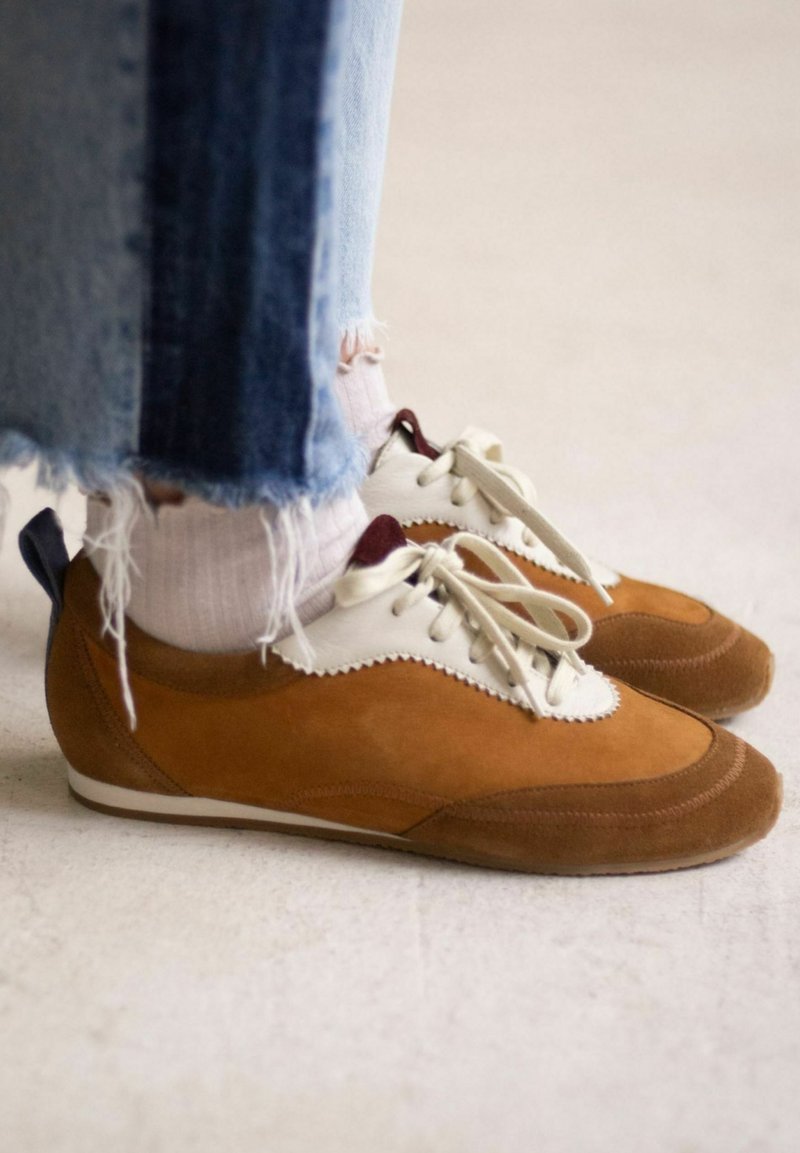 Scarpe in suede color sabbia con dettagli in pelle bianca, di forma arrotondata e con lacci. Jeans blu scoloriti e calzini bianchi con righe leggere visibili.