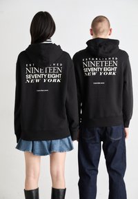 Juodos sportinės hoodies su baltais užrašais „ĮSTEIGTA DEVYNI ŠIMTAI Aštuoni NEW YORK“ ant nugaros, derinamos su džinsiniu sijonu ir tamsiomis kelnėmis.